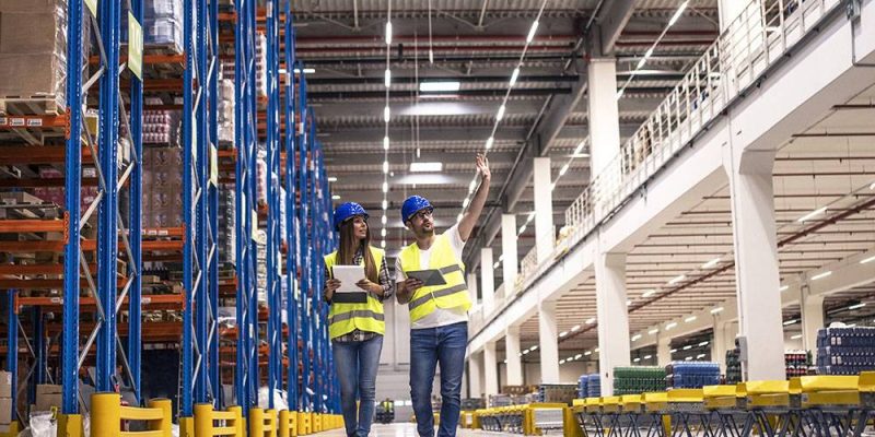 Distribution warehouse interior with workers wearing hardhats and reflective jackets walking in storage area.